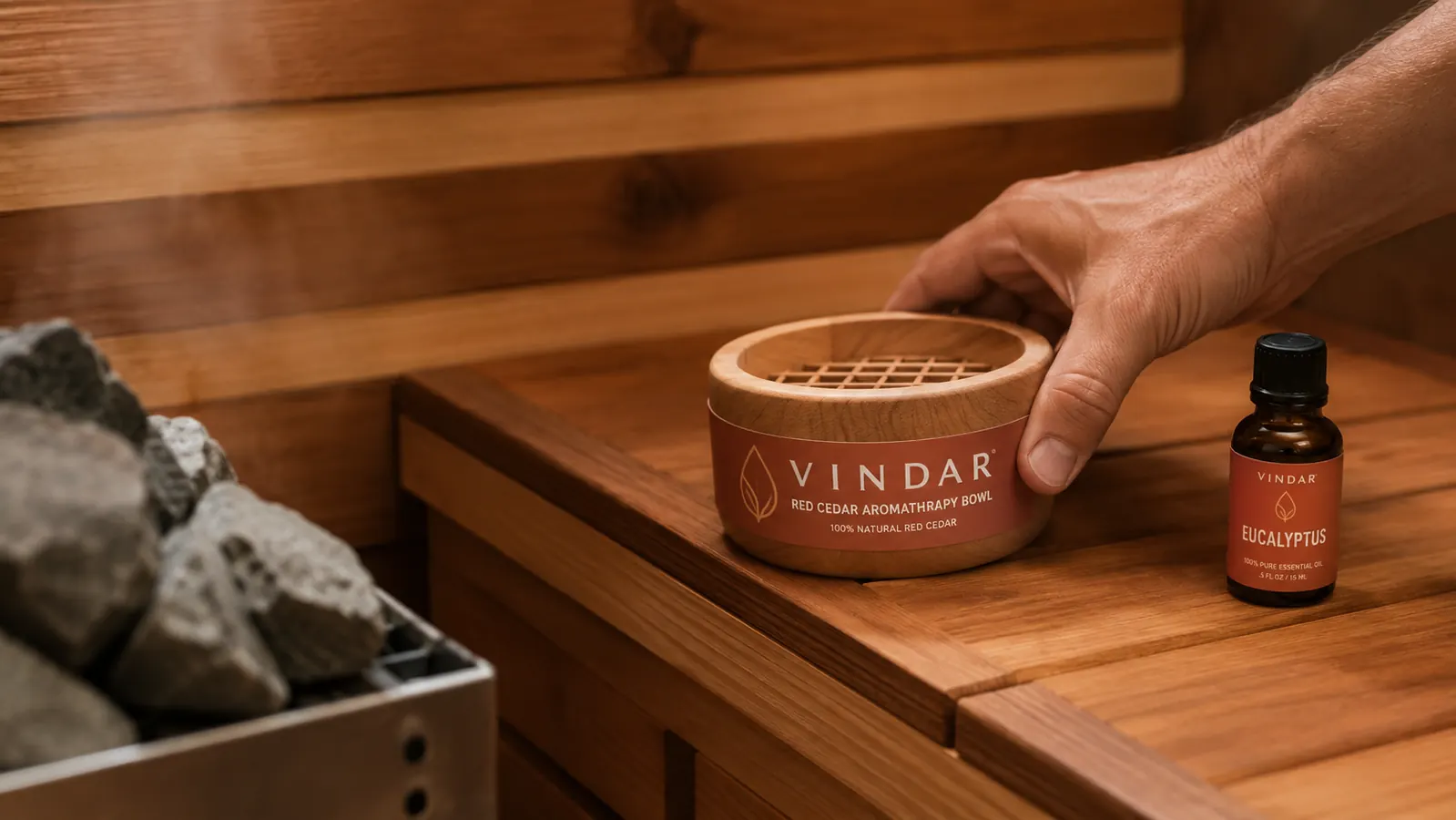 Hand placing a red cedar aromatherapy bowl on the corner of a sauna bench near the heater rocks, oil bottle beside it
